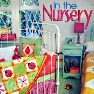 May include: A nursery scene featuring a white metal crib with a colorful quilt adorned with ladybugs and flowers. The quilt is paired with a pink and green crib skirt. The image includes the text "In the Nursery" and the names Jennifer Sampou & Carolyn Schmitz.