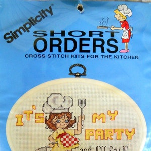 May include: Cross-stitch kit titled "It's My Party" featuring a cartoon chef holding a spatula and a checkered apron. The kit includes a 7" x 5" oval frame, chart, needle, instructions, and cotton floss. The packaging is blue.