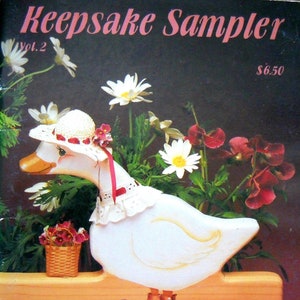 May include: A painted wooden goose wearing a hat and a lace collar, standing on a wooden base with two pegs. The image is from a book titled "Keepsake Sampler Vol. 2" by Charleen Stempel & Sue Schecwe. The background features flowers.