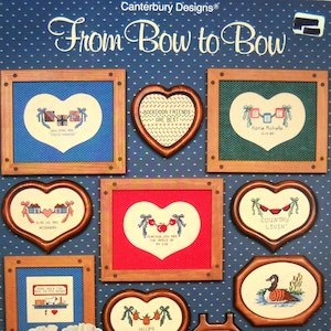 May include: A collection of framed cross-stitch samplers with various heart and house shapes. The samplers feature embroidered text and images, including phrases like "Backdoor Friends Are Best" and "Welcome to Our Neighbors." The color palette includes blue, red, and green.