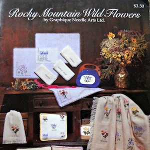 May include: A collection of embroidered items featuring Rocky Mountain wildflowers. The image includes a book, pillows, towels, and a blanket, all showcasing floral designs. The color palette includes beige, white, and various floral colors.