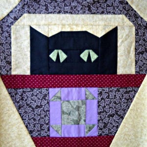 May include: A square quilt block featuring a stylized black cat face with light green triangular eyes. The cat is framed by cream-colored borders and dark purple fabric with a white leaf pattern. A central square block features purple and gray.