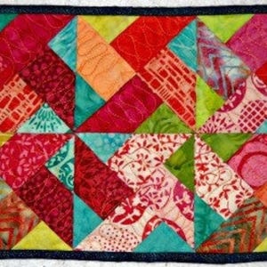 Op de afbeelding: Een rechthoekige, handgemaakte quilt met een levendig patchwork-ontwerp. De quilt toont een verscheidenheid aan kleurrijke stoffen in tinten rood, roze, oranje, groen en blauw, gerangschikt in een geometrisch patroon. De quilt is omzoomd met een donkerblauwe stof.