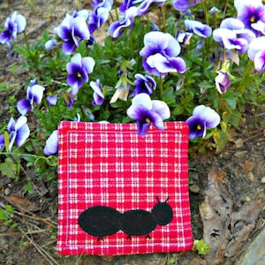 May include: A red and white checkered fabric coaster with a black ant applique. The coaster is set in front of a bed of purple and white pansies. The coaster is square and has a stitched edge.