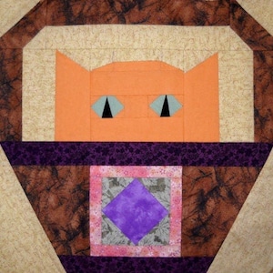 May include: A square quilt block featuring a stylized orange cat face with triangular ears and light blue eyes with black pupils. The cat is set against a brown marbled background with a central purple diamond patch framed by pink floral fabric.