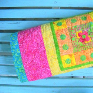 May include: A colorful, handmade quilt with a vibrant floral design. The quilt features squares of pink, yellow, orange, and green fabric, with a large pink flower applique. The quilt is quilted with yellow thread and has a blue and green border.