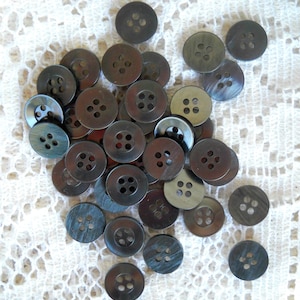 May include: A collection of dark gray, round buttons with four holes each. The buttons have a slightly iridescent sheen and are scattered on a white, open-weave fabric. Ideal for sewing and craft projects.
