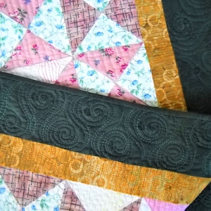 May include: Close-up of a handmade quilt with a patchwork design. The quilt features triangles of pink, white, and floral fabrics. A dark green border with a swirling pattern and a gold border with a floral pattern are also visible.