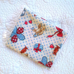 May include: A folded piece of fabric featuring a cream background with a repeating pattern of brown teddy bears, red hearts, blue and red plaid flowers, wrapped gift boxes with red bows, and red candies scattered across the surface.
