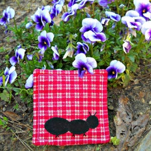 May include: A red and white checkered fabric coaster with a black ant applique. The coaster is surrounded by purple and white pansies and green foliage. The coaster is square-shaped.
