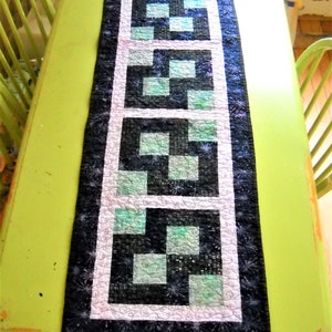 May include: A rectangular quilted table runner with a repeating geometric pattern. The design features black and teal squares framed by white and a dark blue border. The runner is placed on a lime green table.