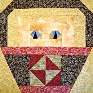 May include: A geometric quilt block featuring a cat's face. The cat has blue eyes and is set against a background of yellow, brown, and red fabrics. The design includes various shapes and patterns, creating a patchwork effect.