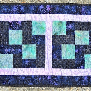 May include: A rectangular quilted table runner featuring four square blocks with a geometric pattern in shades of teal, black, and purple. The border is a dark fabric with a purple starburst print, framed by a light lavender inner border.