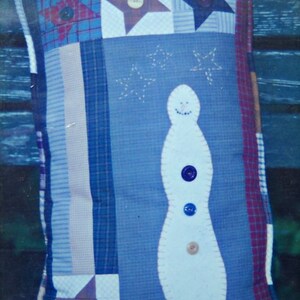 May include: A decorative pillow with a patchwork design featuring a snowman and star motifs. The pillow has a blue background with a white snowman applique and is adorned with various buttons. The edges are trimmed with a plaid fabric.