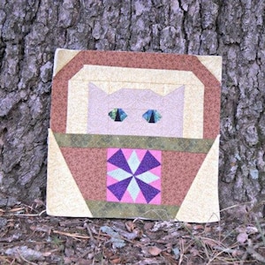 May include: A handmade quilt block featuring a cat design. The cat's face is light pink with blue eyes. The block has a brown and tan border, and a central square with a starburst pattern in pink, purple, and green.