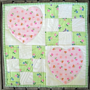 May include: A handmade patchwork quilt with a green and white gingham border. The quilt features two pink gingham hearts with floral patterns and green and white checkered squares with floral accents. The quilt is a charming, decorative item.