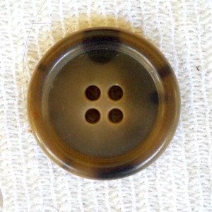 May include: A large, round button with four holes. The button is a mix of brown and tan colors, with a darker rim and a lighter center. The button is set against a white, textured background.