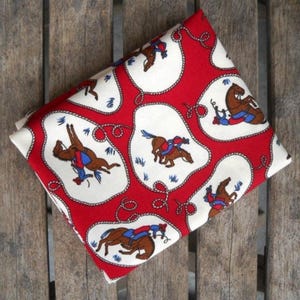 May include: Folded red fabric with a repeating pattern of white ovals containing brown horses and riders. The ovals are outlined with a white rope design. The background is a wooden surface.