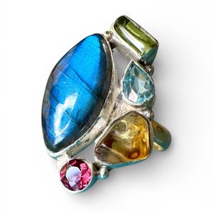 May include: A silver ring featuring a large marquise-cut blue labradorite stone as the centerpiece, accented by four smaller faceted gemstones in shades of green, light blue, yellow, and pink, all set in a polished metal band arrangement.