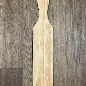 May include: A plain wooden paddle with a rounded top and a hole in the center. The paddle is light brown with a natural wood grain.