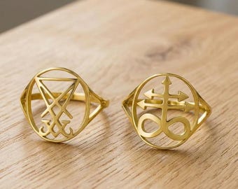 May include: Two gold-colored rings with intricate designs. One ring features a symbol within a circle, while the other displays a cross-like emblem with arrows. The rings are set against a wooden surface.