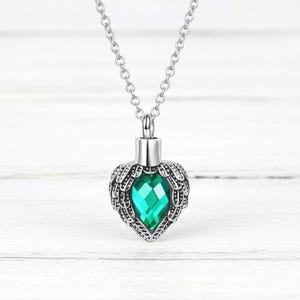 May include: A silver-toned necklace with a heart-shaped pendant. The pendant features a faceted emerald-green gemstone set within a detailed wing design. The necklace hangs against a white wooden background.