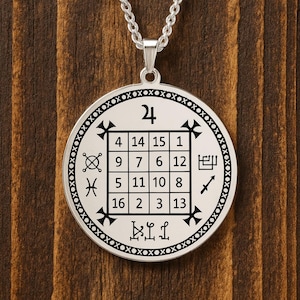 May include: A silver-colored pendant necklace with a circular charm featuring a grid of numbers and symbols. The pendant hangs from a chain and is set against a wooden background. The charm has a diameter of approximately 1.5 inches.
