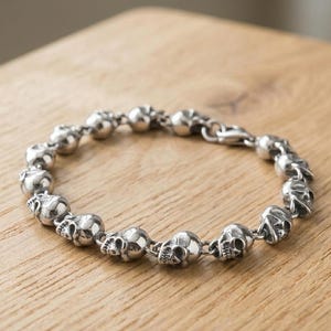 May include: A silver bracelet featuring a series of skull charms. The bracelet is made of metal and has a clasp closure. The skulls are detailed and evenly spaced along the chain. The bracelet is displayed on a wooden surface.