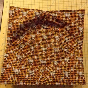 May include: A square fabric bowl cozy with a repeating pattern of small circles in shades of brown, orange, and white. The cozy is folded to create a bowl shape, with visible seams and folds.