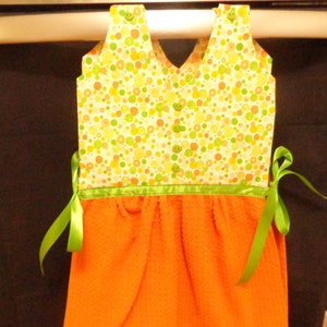 May include: A green and orange kitchen towel with a white fabric top with a green and yellow polka dot pattern. The towel has green ribbon ties.