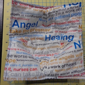 May include: White, square bowl cosy with a repeating text pattern in various colours. The text includes phrases like "Nurses are patient people" and "Healing". The word "Angel" is in blue.