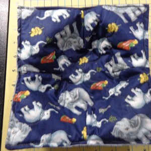May include: A navy blue fabric bowl cosy with a repeating pattern of white elephants, yellow flowers, and red carrots. The cosy is quilted and designed to hold a bowl of hot food. The edges are neatly sewn.