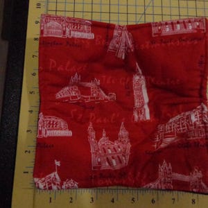 May include: Red fabric coaster with white outlines of London landmarks, including Buckingham Palace, St. Paul's Cathedral, and the Tower Bridge. The text "Buckingham Palace", "St. Paul's", and "The Tower Bridge" are printed on the coaster.