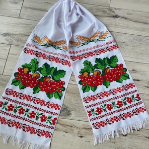 May include: White embroidered scarf with red berries, green leaves, and wheat stalks. The scarf has a fringed edge and intricate floral patterns in red and green. The design is symmetrical, with the same pattern on both ends.
