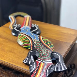 May include: A wooden lizard sculpture featuring intricate dot art patterns in vibrant red, yellow, green, and blue colours. The lizard has a black base colour with detailed white dot accents on its limbs and a curved tail, showcasing a textured finish.