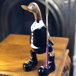 May include: A wooden duck figurine featuring a natural brown head and a black tuxedo-style body with a white shirt front, three yellow buttons, and a black bowtie. The duck wears black boots with white laces and holds a thin purple stick.