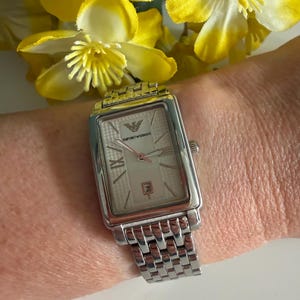 May include: Silver rectangular wristwatch with a light-coloured face, Roman numeral hour markers, and a date display. The watch features a silver link band and the Emporio Armani logo. The watch is shown on a wrist with yellow flowers in the background.