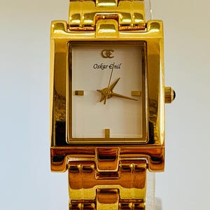 May include: A gold-coloured wristwatch with a rectangular face and a matching link strap. The watch face is white with gold hands and the text "Oskar Emil". The watch has a small black crown.