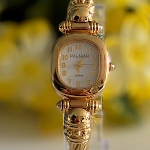 May include: A gold-tone quartz watch with a cushion-shaped case and a white dial featuring the text Joan Rivers Classics. The watch has a polished gold-tone bezel and a textured cable-style band with decorative gold-tone metal accents at the lugs.