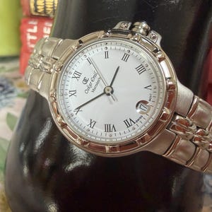 May include: A silver Oskar Emil Marrakech watch with a white face, black hands, and Roman numeral hour markers. The watch has a date display at the 3 o'clock position and a silver link band.