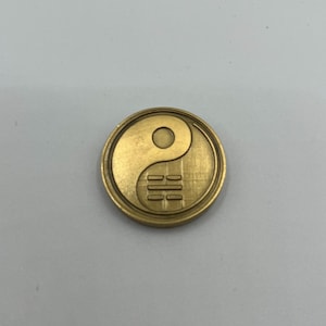 Könnte beinhalten: Eine runde, goldfarbene Münze mit einem Yin-und-Yang-Symbol. Die Münze hat ein erhabenes Design und eine strukturierte Oberfläche. Das Yin-und-Yang-Symbol ist eine klassische Darstellung von Ausgewogenheit und Harmonie.