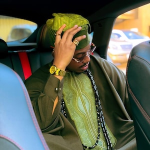 May include: An olive green outfit, including a hat and tunic with detailed embroidery, and a beaded necklace. Gold watch and rings complete the look. The person is seated in a car. The outfit is a traditional style.