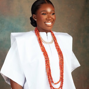 Peut inclure: Une femme porte un haut blanc texturé et un collier de perles de corail orange superposées avec des accents dorés. Un collier en chaîne dorée est également visible. La femme a les cheveux attachés et sourit.