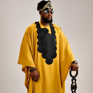 Puede incluir: Un atuendo africano tradicional amarillo mostaza con bordados negros y un tocado a juego. El atuendo incluye una túnica larga y se combina con un bastón negro y gafas de sol.