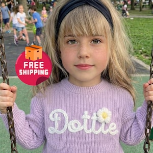 Op de afbeelding: Een jong persoon draagt een lavendelkleurige gebreide trui met de naam "Dottie" in witte letters en een witte madeliefjesapplicatie. De persoon draagt ook een zwarte hoofdband en zit op een schommel.