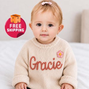 May include: A cream-colored baby sweater with the name "Gracie" embroidered in a reddish-orange script. A pink flower is stitched on the right side. The sweater is worn by a baby with a pink bow in their hair.