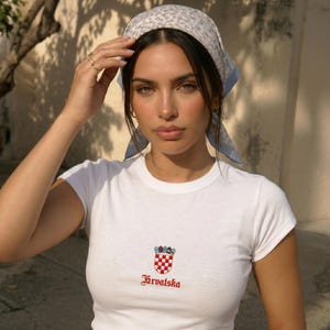 Puede incluir: Una camiseta blanca de manga corta con un pequeño escudo croata bordado en cuadros rojos y blancos con una corona encima, y la palabra Hrvatska en texto rojo debajo. Se lleva un pañuelo para la cabeza con estampado floral azul claro y blanco.