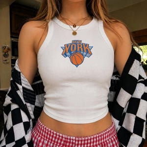 Puede incluir: Una camiseta de tirantes blanca acanalada con un estampado gráfico del texto NEW YORK en letras azules y naranjas sobre un icono de baloncesto naranja. La prenda se lleva con una rebeca de cuadros blancos y negros y un pantalón de cuadros rojos y blancos.