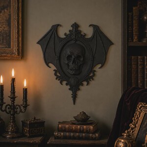 Peut inclure: Une plaque décorative murale noire présentant un crâne humain central encastré dans un cadre ovale. Le cadre est orné de volutes complexes et de grandes ailes de chauve-souris symétriques s'étendant sur les côtés, créant un relief sombre.