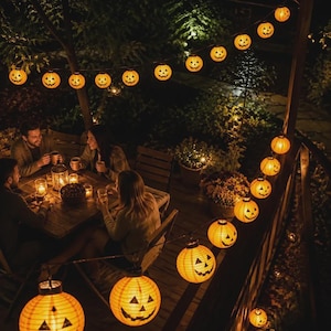 May include: A string of orange jack-o'-lantern paper lanterns illuminated with warm light, hanging in a curved line across a wooden deck. Each round lantern features a black printed face with triangular eyes and a jagged mouth design.